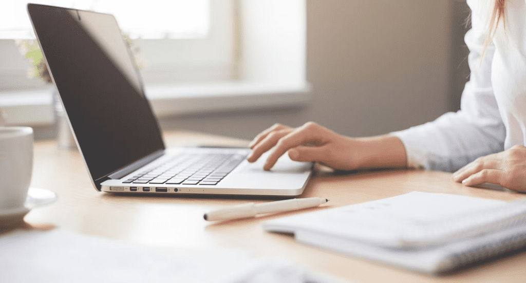 A woman using a laptop on an office table with coffee, pen and notebook.