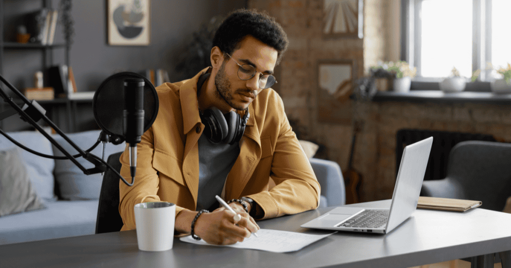 A man in a yellow jacket is writing in a notebook at a desk with a microphone, concentrating deeply on his work; a sharp, specific focus is key to standing out from the crowd.