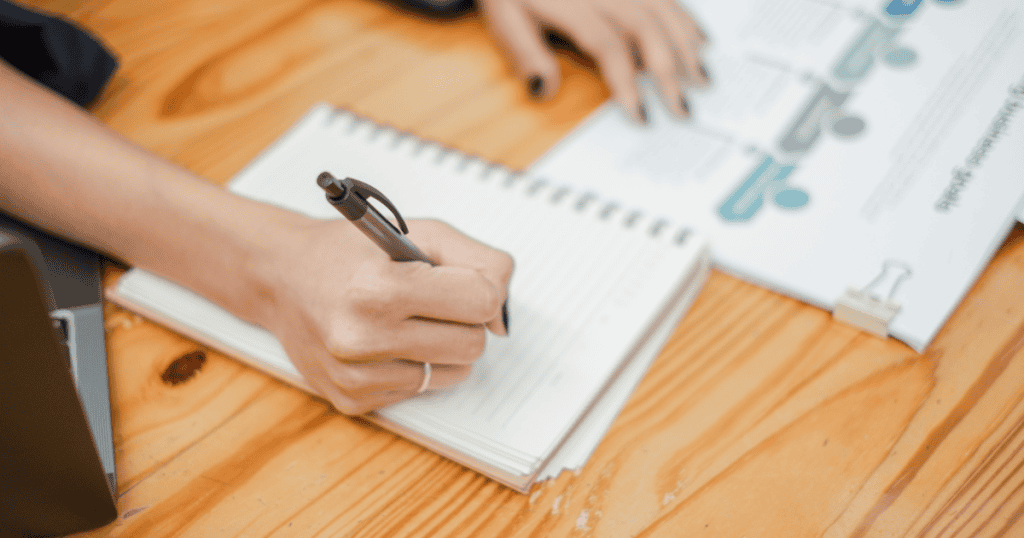 A top-down view of someone writing in a notebook next to an organized document, showcasing the methodical process behind generating consistent output.