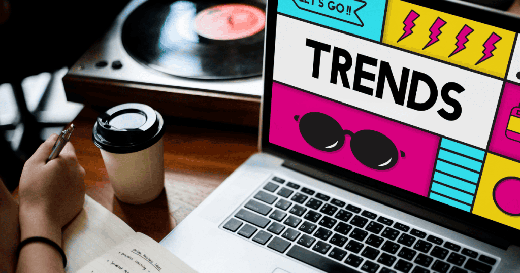 A person takes notes beside a laptop displaying a vibrant pop-art trends graphic, actively researching current market patterns to discover under-served topics to write about.