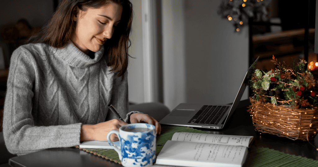 A woman smiles while writing in a notebook next to her laptop, illustrating the focused dedication required to produce meaningful work regularly.