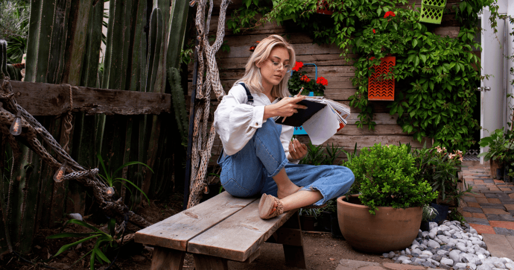 A woman in overalls sits on a garden bench surrounded by lush plants and cacti while reviewing her notebook, finding inspiration for cultivating content rooted in nature and exterior design.