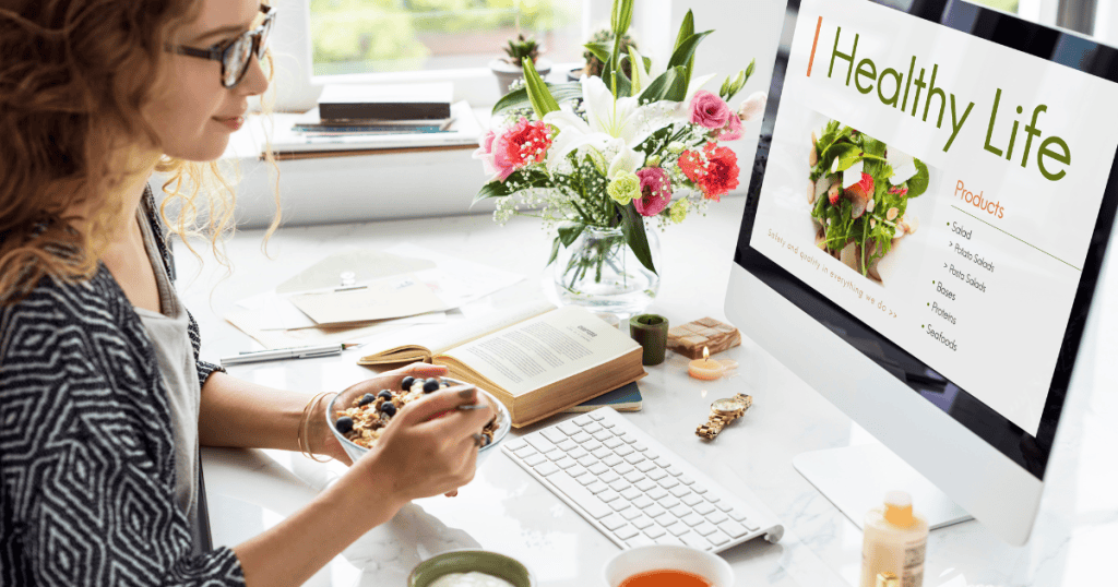 A woman enjoys a nutritious bowl of fruit while browsing a website featuring fresh salad recipes on her desktop, highlighting the variety of topics available within the lifestyle and physical well-being sector.