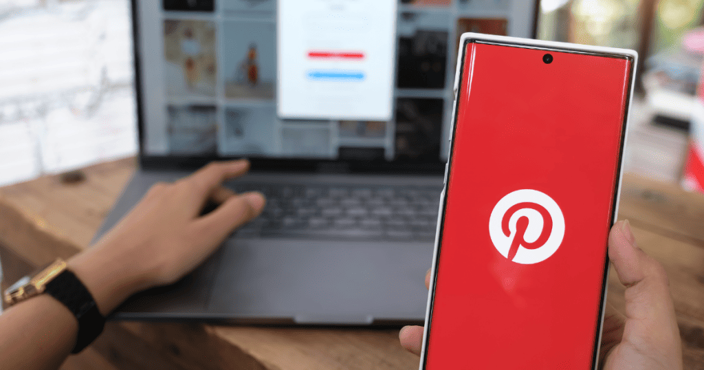 A close-up shows a hand holding a smartphone with the red Pinterest logo against a blurred laptop background, visually linking the social media app’s usage to the website content displayed behind it.