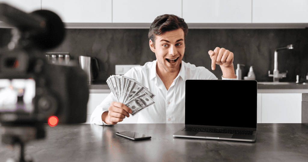 A smiling man holds a fan of cash while pointing toward a laptop screen, illustrating the potential for generating income through digital content creation.