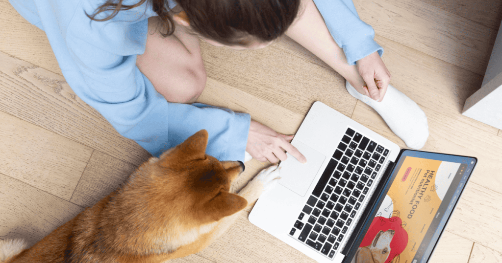 A woman sits on the floor with her dog while browsing a pet nutritionist website on her laptop, demonstrating the specific online resources available for dedicated animal owners.
