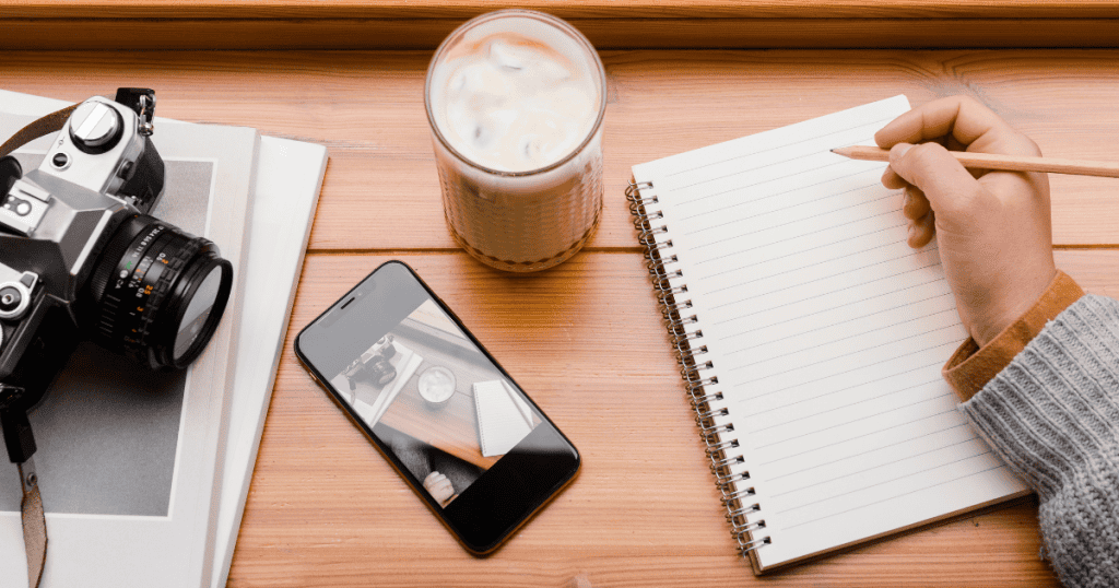 A vintage camera sits beside a smartphone and iced coffee on a wooden desk as a person writes notes, illustrating the process of narrowing down specific visual subjects to capture.