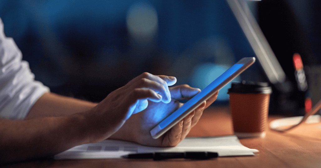 A person's hands navigate a glowing tablet in a dimly lit room, emphasizing the use of modern mobile interfaces for staying connected after hours.