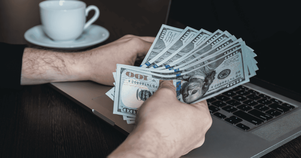 A person holds a fanned-out stack of hundred-dollar bills over a laptop keyboard, representing the financial rewards that remain out of reach when common digital publishing errors persist.