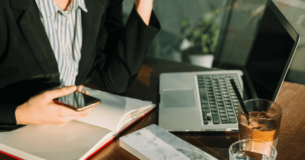 A professional woman looks at her phone while sitting at a desk with a laptop and coffee, capturing the busy atmosphere of managing a project's foundational requirements.