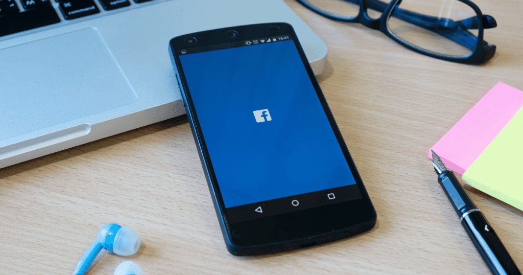 A smartphone displaying the Facebook logo sits on a wooden desk alongside office supplies, representing the everyday tools where subtle oversights often occur.