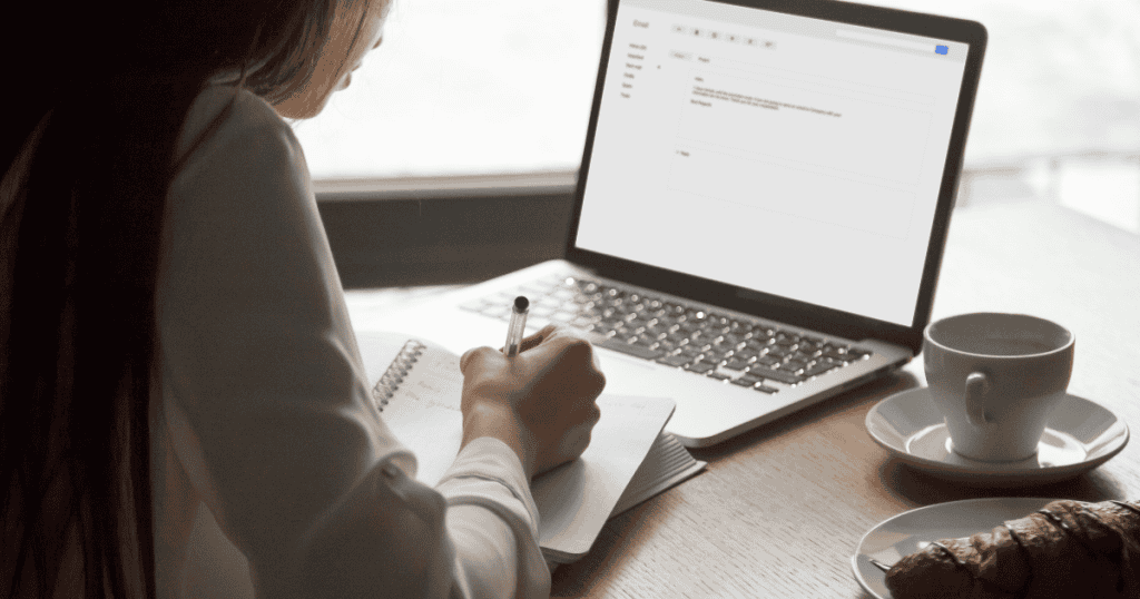 A woman writes in a notebook beside a laptop displaying an open email interface, showcasing the focused effort required to manage a direct communication channel with followers.