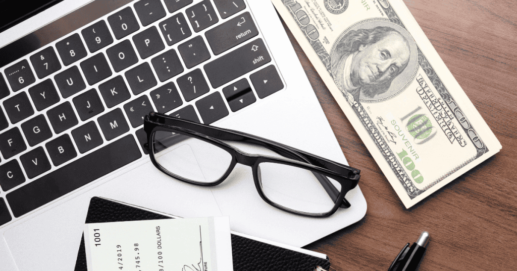 A top-down view of a laptop next to a stack of hundred-dollar bills and a check, hinting at the logistical complexities of managing digital revenue.