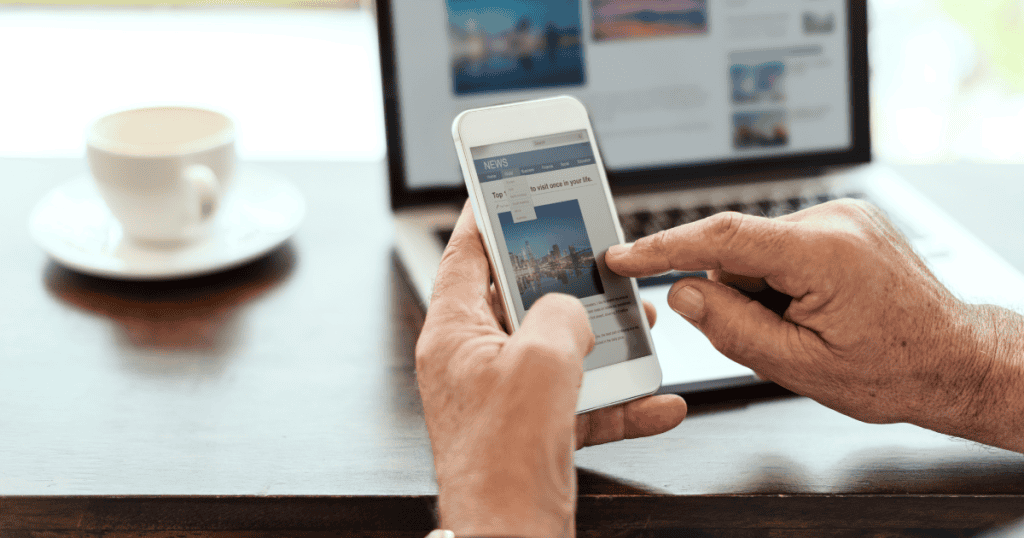 A hand interacts with a news app on a smartphone while a laptop remains open in the background, mirroring the flow of information across different devices.