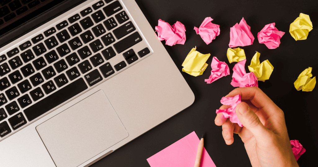 A hand reaching for crumpled paper scraps next to an open laptop, symbolizing the trial-and-error nature of the creative journey for new writers.