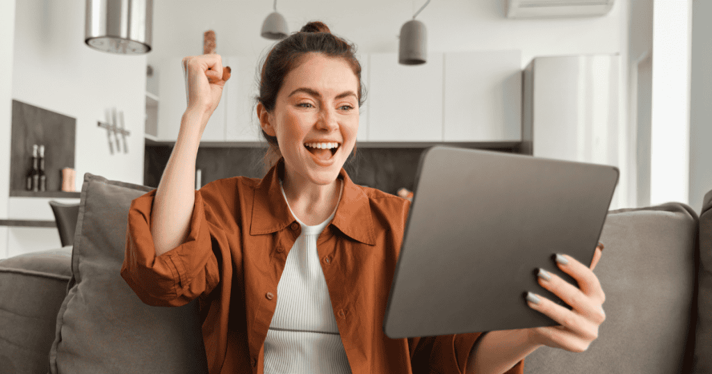 A woman cheers with a raised fist while looking at her tablet in a bright living room, capturing the genuine excitement of finding a community that resonates.